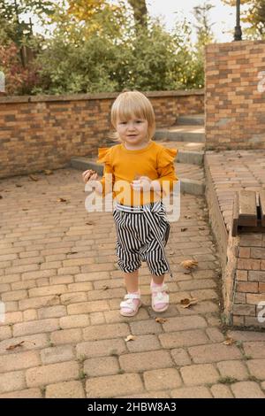 A beautiful cheerful little girl in a stylish striped autumn suit plays with autumn leaves in a city park. Lifestyle Stock Photo