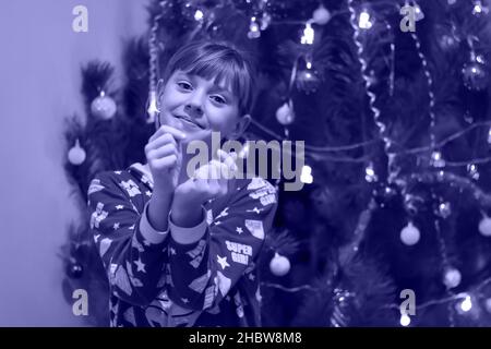 Defocus little blonde Caucasian girl 10 years old in pajamas stands near the Christmas tree. Shows Korean heart sign, love symbol. Color of the year Stock Photo