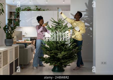 Afro woman taking photo of Christmas firtree on smartphone, african couple set up live New Year tree Stock Photo