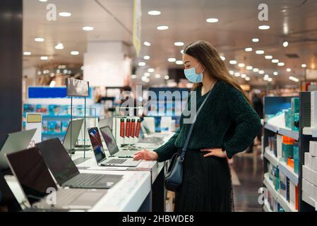 Focused young woman in face mask examines computers on display in tech store department trying to choose laptop for remote work online during Covid19 outbreak. Shopping upon pandemic time concept Stock Photo