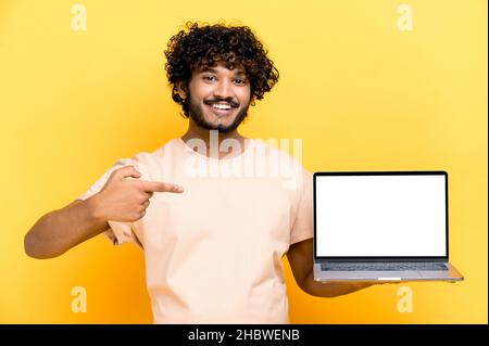 Satisfied positive modern indian curly-haired guy, holds an open laptop with blank white screen, points finger at it, standing on isolated orange background, smiling friendly. Mock-up, copy space Stock Photo