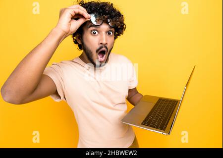 Amazed happy excited Indian or Arabian guy, in casual shirt, take off glasses, holds an open laptop in hand, looks surprised and happily at the camera while standing on isolated orange background Stock Photo