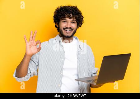Positive handsome indian or arabian curly-haired guy, student, freelancer, holds an open laptop, points OKAY gesture, standing on isolated orange background, smiling friendly. Mock-up, copy space Stock Photo