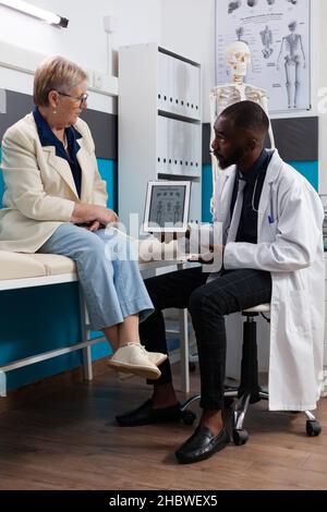 Specialist therapist doctor holding tablet showing body radiography explaining bones pain discussing healthcare treatment during medical appointment in hospital office. Senior woman patient on bed Stock Photo