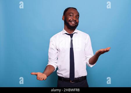 Portrait of smiling businessman with open palms weighing options on blue background. Confident office worker doing hand gesture comparing pros and cons putting decision in balance. Stock Photo