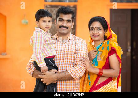 Portrait of young rural indian family standing outdoor at village house. Parent holding their little child, Mustache man wearing kurta and woman wear