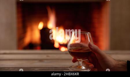 Drink alcohol and relax, winter time. Man holding a brandy glass, wooden table and burning fireplace background, space. Warm cozy home interior Stock Photo
