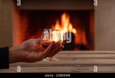 Man holding a brandy glass, wooden table and burning fireplace background, space. Drink alcohol and relax, winter time. Warm cozy home interior Stock Photo