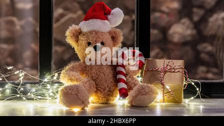 Wait for Santa. Teddy bear sitting on a window sill, Christmas lights and gift box, warm festive home interior Stock Photo