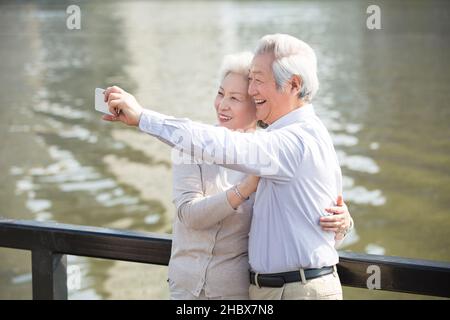 Happy elderly couple taking selfies on their mobile phone by a lake Stock Photo