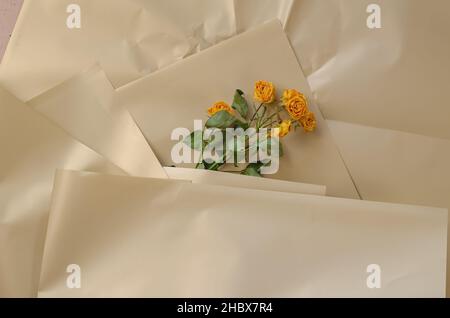 Wilted flowers against a creamy surface. Yellow roses. Crumpled shiny wrapping paper. Top view. Selective focus. Stock Photo
