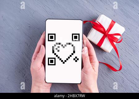 Hands holding mobile phone with white screen with heart shaped qr code and gift box with red ribbon on gray background. Online shopping for valentine' Stock Photo