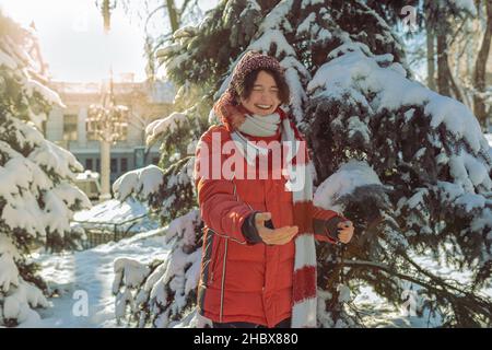 Cheerful laughing teenage girl in winter warm clothes throws snow and plays in winter park in sunny frosty weather Stock Photo
