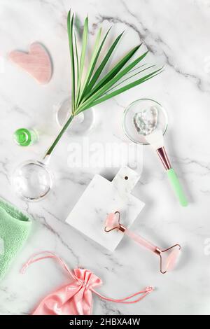 Facial massage. Moisturizer, pink rosa quarz face roller and gua sha stone on marble surface. Green plant leaf is Cylindric Sedge. Stock Photo
