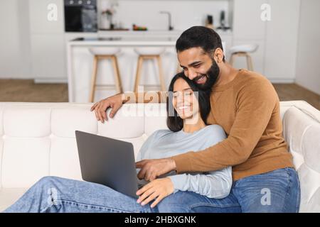 Young Pakistani man pointing at the screen while using laptop on the sofa at home with his wife. Spouses watching something on laptop. Happy man and woman embracing and relaxing Stock Photo