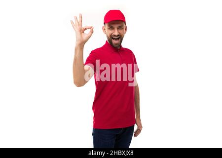 courier in red clothes on a white background shows a gesture of the symbol Okay, looks at the camera with a smile Stock Photo