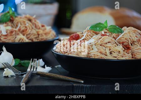 Selective focus of two plates of vegetarian spaghetti pasta with fresh basil leaves and parmesan cheese over a black rustic wood table with blurred fo Stock Photo