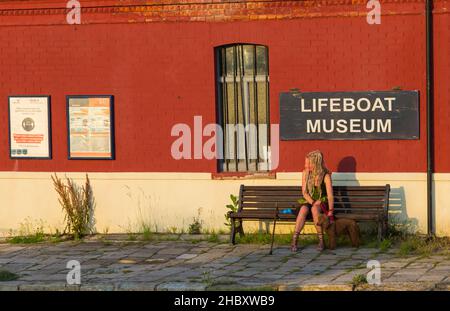 Woman sitting on bench with dog enjoying the evening sunshine outside Lifeboat Museum at Poole, Dorset UK in July Stock Photo