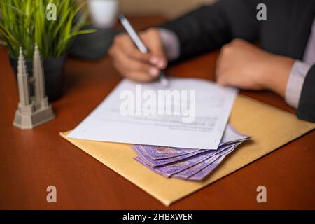 Corrupt Businessman or CEO or Politician hands holding a pen while signing documents or contracts with money under it on an office desk. Stock Photo