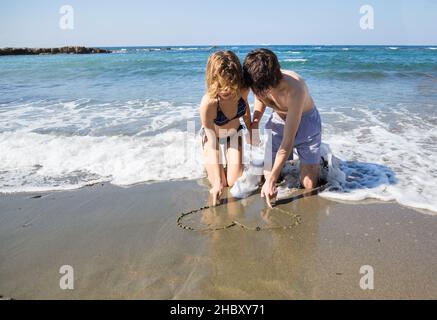 young woman and man in love, draw a heart on the sand while sitting on the seashore. Valentine's day on vacation, first love, happy relationships, sum Stock Photo