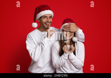 Cheerful Man In Santa Hat Covering Girlfriend's Eyes And Showing Shh Sign Stock Photo