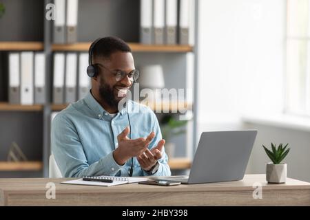 Happy millennial african american bearded man in glasses and headphones gestures and works on laptop Stock Photo