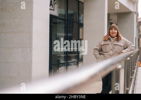 Happy smiling overweight woman in warm hat and jacket standing posing near railing of office building at city street in cloudy autumn day. Stock Photo