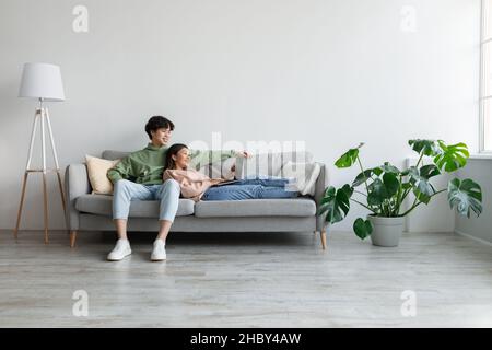 Cheery young Asian couple sitting on sofa, using laptop computer, communicating remotely, having video call at home Stock Photo