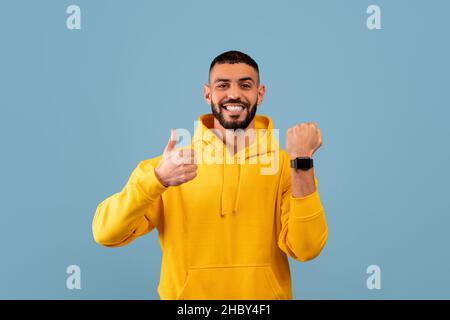 Handsome young arab man wearing smart watch and showing thumb up, recommending modern gadget, blue background Stock Photo
