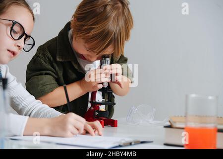 Cringe little boy looking through the microscope, girl taking notes next to him Stock Photo