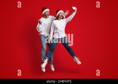 Joyful Couple Wearing Santa Hats Jumping And Taking Selfie With Smartphone Stock Photo