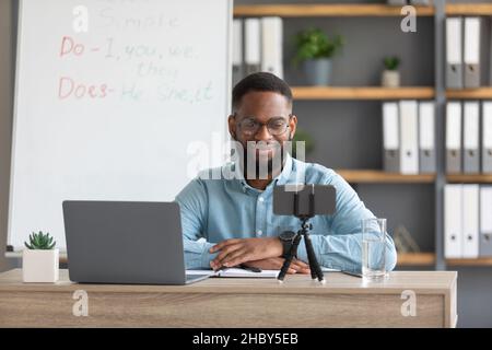 Smiling young handsome african american man teacher in glasses looks at laptop, phone webcam in home Stock Photo
