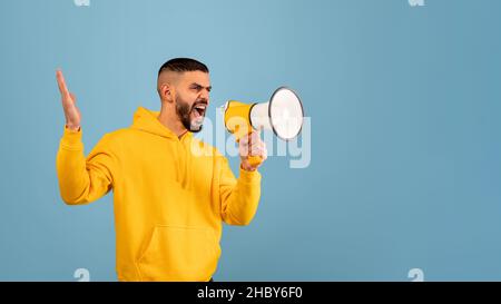 Making important announcement. Angry young arab man shouting using megaphone, blue background, copy space Stock Photo