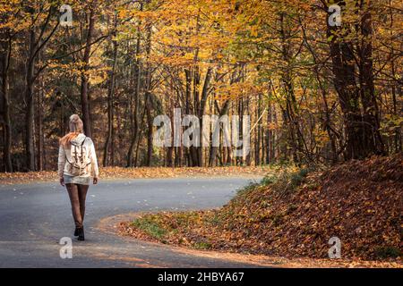 Woman walking on the road in the middle of forest at autumn season. Traveler with backpack on empty country road. Stock Photo
