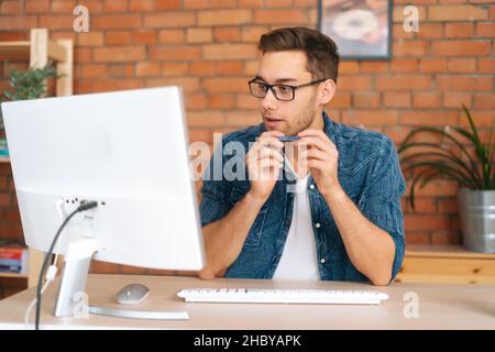 Portrait of thoughtful handsome young freelance programmer male in stylish glasses working on desktop computer holding pen in hands Stock Photo