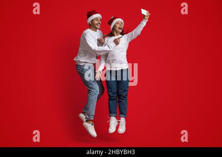 Positive Man And Woman Wearing Santa Hats Jumping And Taking Selfie Together Stock Photo