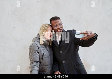 Diverse boyfriend and girlfriend in outerwear smiling and taking selfie against gray wall during date on city street Stock Photo