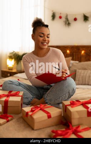 Excited black lady writing greeting card for friend, wrapping gift boxes and smiling, sitting on bed at home Stock Photo