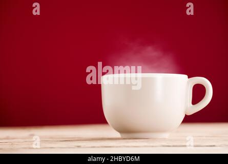 broken white cup of tea or coffee with hot steam smoke on rustic wooden table, red background Stock Photo