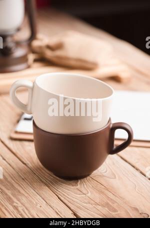 Two cups of coffee or tea stacked on old wooden table, with notebook, raffia sack, lamp Stock Photo