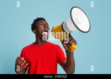 Portrait Of Emotional African American Guy Shouting With Megaphone Over Blue Background Stock Photo
