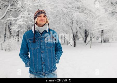 Portrait of young man in snowy winter park wearing jeans coat funny hat walking outdoors. Guy enjoys falling snow Stock Photo