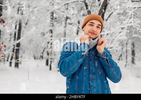 Portrait of stylish man in snowy winter park wearing jeans coat hat posing outdoors. Guy looks at camera Stock Photo