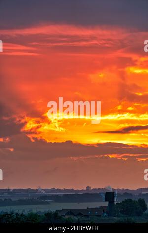 Dramatic orange and yellow sky as the sunrise hidden is by clouds over the South East Kent landscape. A windmill without sails just below the horizon. Stock Photo