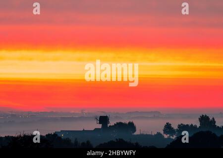 Dawn landscape with a windmill without sails silhouetted against a thick sea mist and above a dramatic sky of red, and yellow bands of cloud. Stock Photo