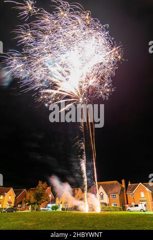 Firework display on village green with fireworks exploding on top of a golden fiery trail in abstract blurry pattern in the sky over a row of houses. Stock Photo