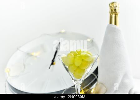 Wine glass with grapes on the background of a blurry clock and a bottle of champagne. Spanish tradition of eating twelve grapes to celebrate New Year. Stock Photo
