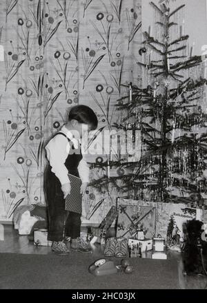Christmas 1962 in Germany. A little boy is amazed and unwraps his presents under the Christmas tree. Stock Photo