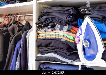Iron and a pile of folded menswear on the shelf. Men's clothing in a closet. Decluttering and cleaning of the home cabinet. Stock Photo
