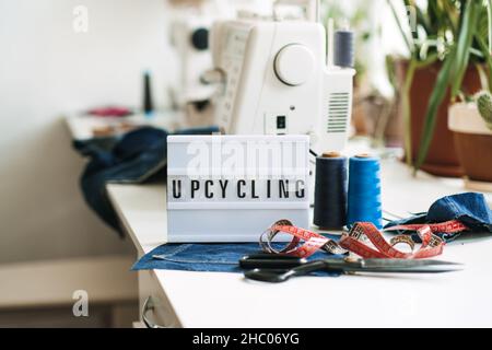 Upcycling text on light board on sewing machines with old blue jeans background. Denim clothes, scissors, thread and sewing tools in sewing studio Stock Photo